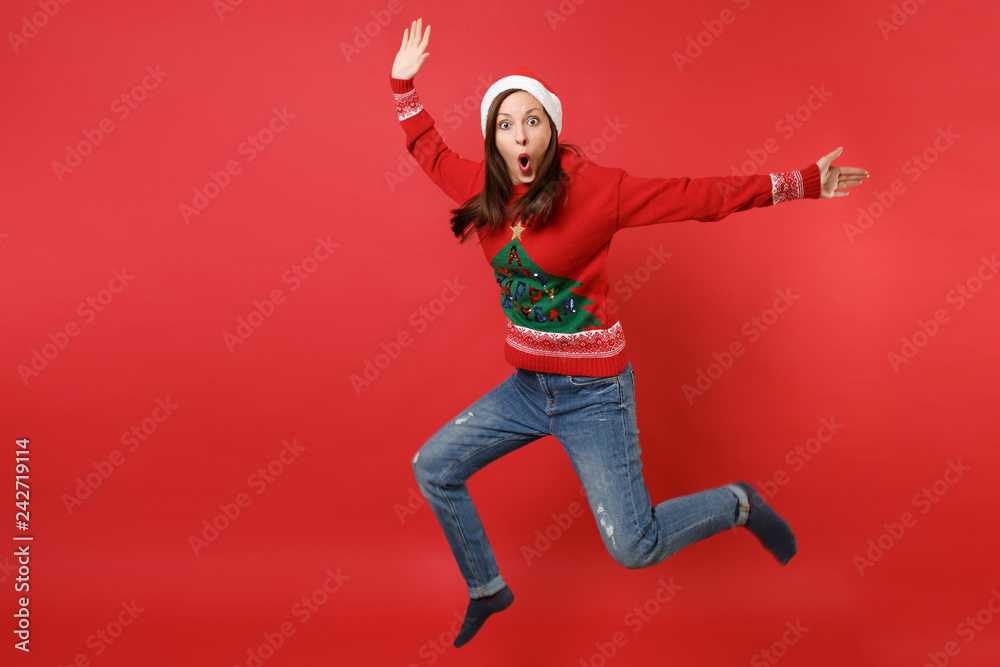 Shocked young Santa girl keeping mouth wide open, looking surprised jumping spreading hands, legs isolated on red background. Happy New Year 2019 celebration holiday party concept. Mock up copy space.