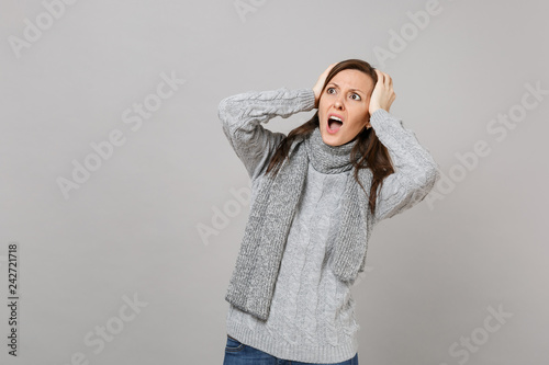 Shocked young woman in gray sweater, scarf looking up, putting hands on head, screaming isolated on grey background. Healthy fashion lifestyle, people emotions cold season concept. Mock up copy space.