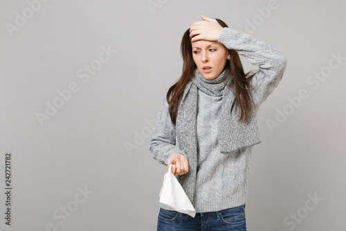 Exhausted woman in gray sweater, scarf hold paper napkin, putting hand on forehead isolated on grey background. Healthy lifestyle, ill sick disease treatment, cold season concept. Mock up copy space.