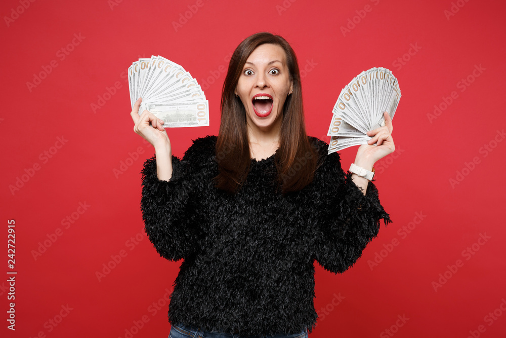 Surprised young woman in fur sweater with opened mouth holding fan of money in dollar banknotes, cash money isolated on red background. People sincere emotions, lifestyle concept. Mock up copy space. Surprised young woman in fur sweater with opened mouth holding fan of money in dollar banknotes, cash money isolated on red background. People sincere emotions, lifestyle concept. Mock up copy space.