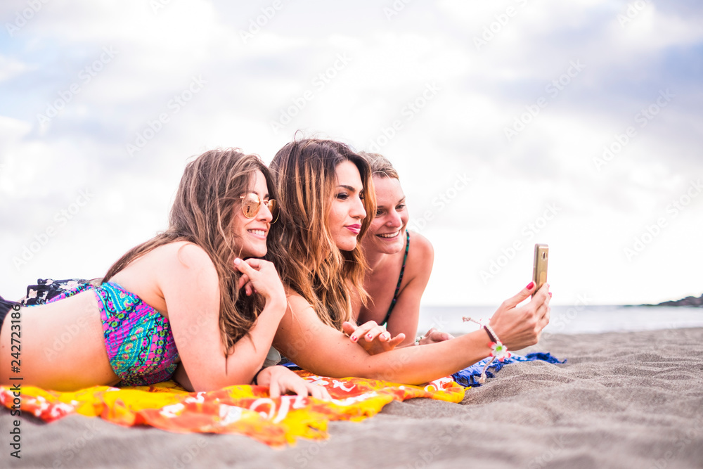 Friendship concept for young millennial girls women caucasian people at the beach under the summer sun playing with phone internet technology taking picture and share it on social to update accounts