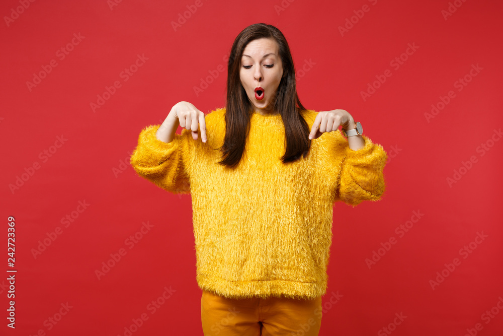 Portrait of shocked young woman in yellow fur sweater pointing index fingers down isolated on bright red wall background in studio. People sincere emotions, lifestyle concept. Mock up copy space. Portrait of shocked young woman in yellow fur sweater pointing index fingers down isolated on bright red wall background in studio. People sincere emotions, lifestyle concept. Mock up copy space.