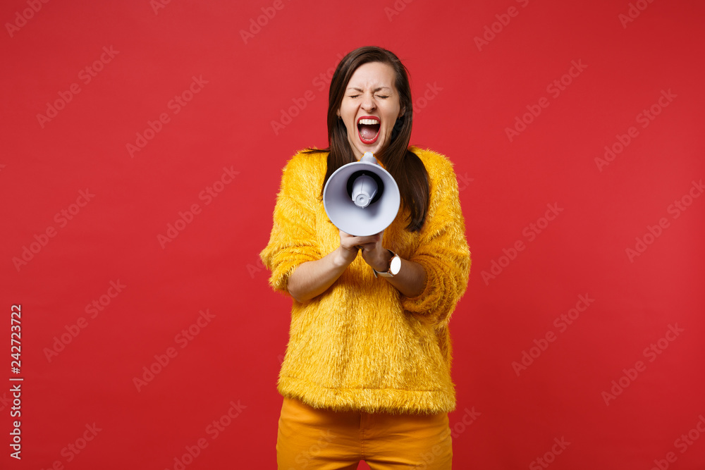 Crazy young woman in yellow fur sweater keeping eyes closed, screaming on megaphone isolated on bright red wall background in studio. People sincere emotions, lifestyle concept. Mock up copy space. Crazy young woman in yellow fur sweater keeping eyes closed, screaming on megaphone isolated on bright red wall background in studio. People sincere emotions, lifestyle concept. Mock up copy space.