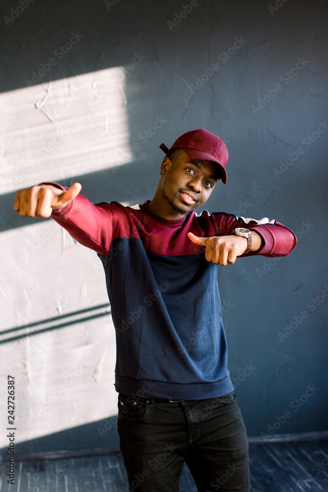 Funny man with dark skin gestures happily feels proud of his success, smiles gladfully, wears old fashionable red cap isolated over dark background. People, happiness and joy concept Funny man with dark skin gestures happily feels proud of his success, smiles gladfully, wears old fashionable red cap isolated over dark background. People, happiness and joy concept