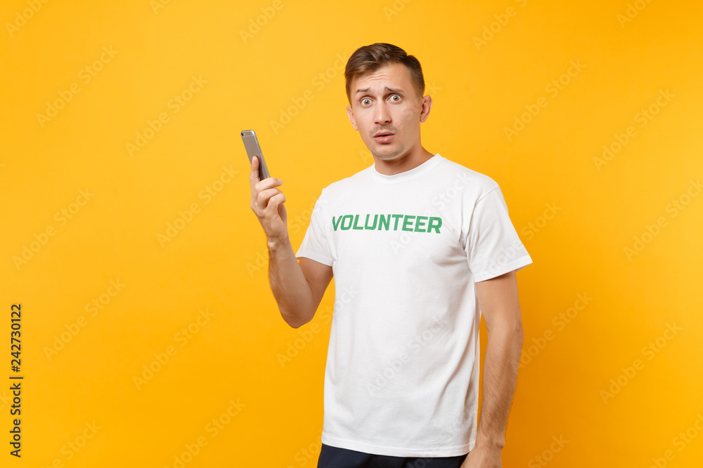 Young man portrait in white t-shirt with written inscription green title volunteer talking on mobile phone isolated on yellow background. Voluntary free assistance help, charity grace work concept. Young man portrait in white t-shirt with written inscription green title volunteer talking on mobile phone isolated on yellow background. Voluntary free assistance help, charity grace work concept.