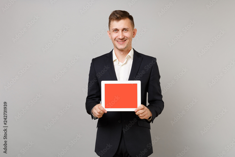 Smiling young business man in classic black suit, shirt holding tablet pc computer with blank empty screen isolated on grey background. Achievement career wealth business concept. Mock up copy space. Smiling young business man in classic black suit, shirt holding tablet pc computer with blank empty screen isolated on grey background. Achievement career wealth business concept. Mock up copy space.