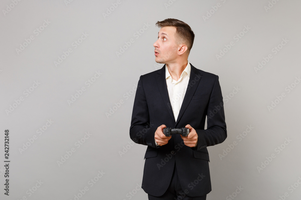 Excited young business man in classic black suit white shirt looking aside playing video game joystick isolated on grey wall background. Achievement career wealth business concept. Mock up copy space. Excited young business man in classic black suit white shirt looking aside playing video game joystick isolated on grey wall background. Achievement career wealth business concept. Mock up copy space.