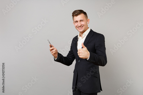 Smiling young business man with wireless earphones listening music, showing thumb up, holding mobile phone isolated on grey background. Achievement career wealth business concept. Mock up copy space.