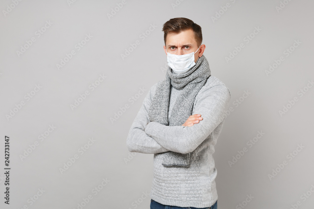 Young man in gray sweater, scarf with sterile face mask holding hands folded isolated on grey wall background. Healthy lifestyle, ill sick disease treatment, cold season concept. Mock up copy space. Young man in gray sweater, scarf with sterile face mask holding hands folded isolated on grey wall background. Healthy lifestyle, ill sick disease treatment, cold season concept. Mock up copy space.