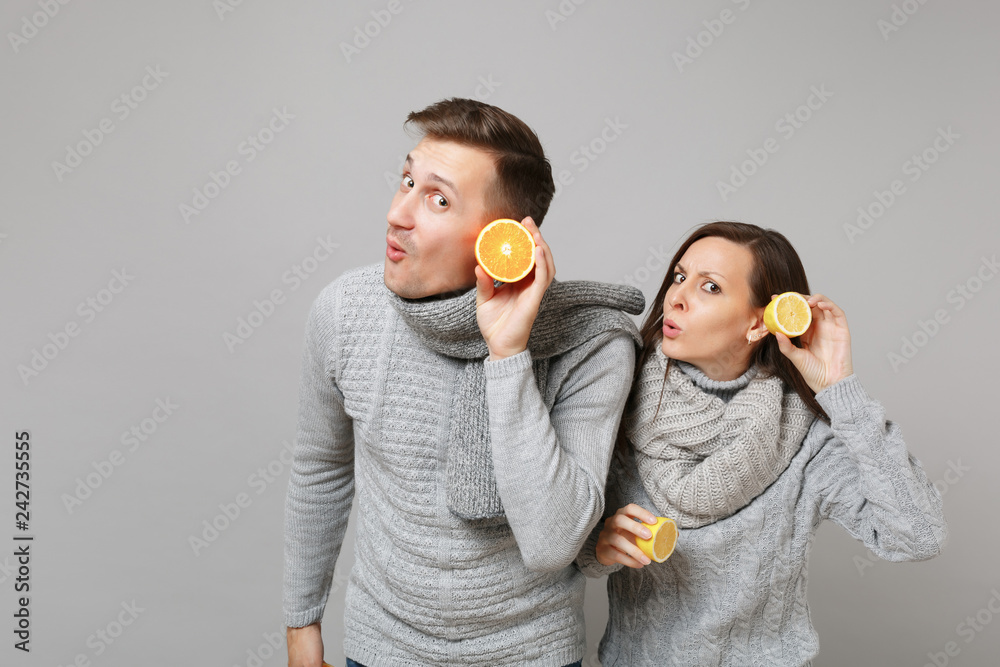 Fun couple girl guy in gray sweaters, scarves together hold orange lemon isolated on grey wall background studio portrait. Healthy lifestyle ill sick treatment cold season concept. Mock up copy space. Fun couple girl guy in gray sweaters, scarves together hold orange lemon isolated on grey wall background studio portrait. Healthy lifestyle ill sick treatment cold season concept. Mock up copy space.