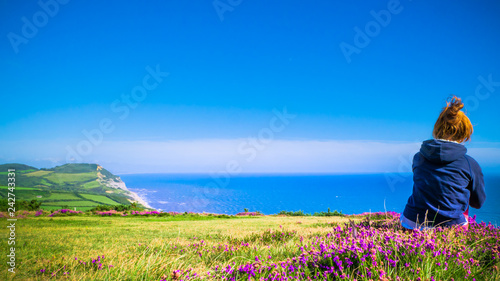 Εκτύπωση καμβά Young redhead girl in trekking clothes is sitting on the top of a hill with purple flowers and sea / English Channel in the front