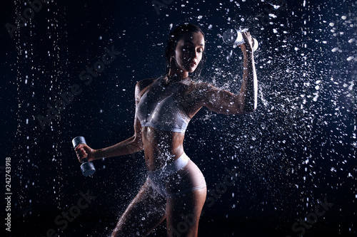 Beautiful young girl in sportswear poses with dumbbells in aqua studio. Drops of water spread about her fitness body. The perfect figure on the background of water splashes