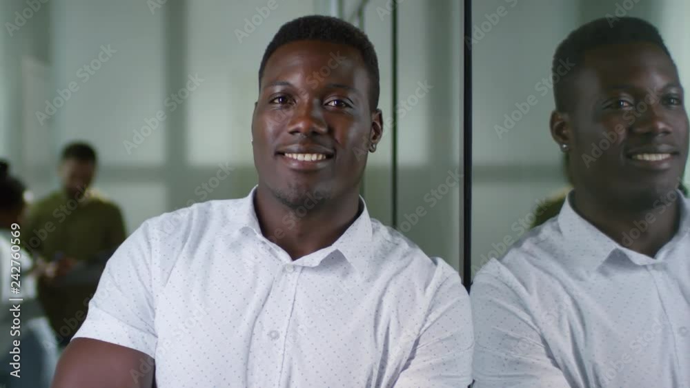 PAN shot of happy black businessman leaning on glass wall in office corridor and smiling for camera. Unrecognizable colleagues discussing work in background PAN shot of happy black businessman leaning on glass wall in office corridor and smiling for camera. Unrecognizable colleagues discussing work in background