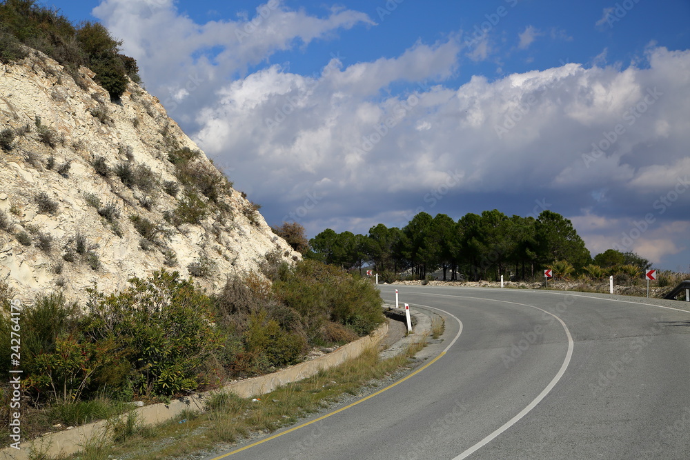 Road in the mountains, curve, sharp turn, arid hill on the left side, trees on the right, cloudy sky