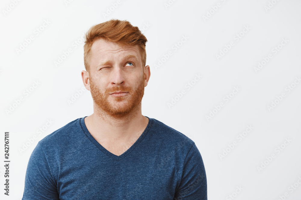 Hmm let us imagine. Portrait of dreamy cute adult redhead male with beard, looking up with one eye closed, lifting eyebrows, thinking or making up idea, picturing something over gray background Hmm let us imagine. Portrait of dreamy cute adult redhead male with beard, looking up with one eye closed, lifting eyebrows, thinking or making up idea, picturing something over gray background