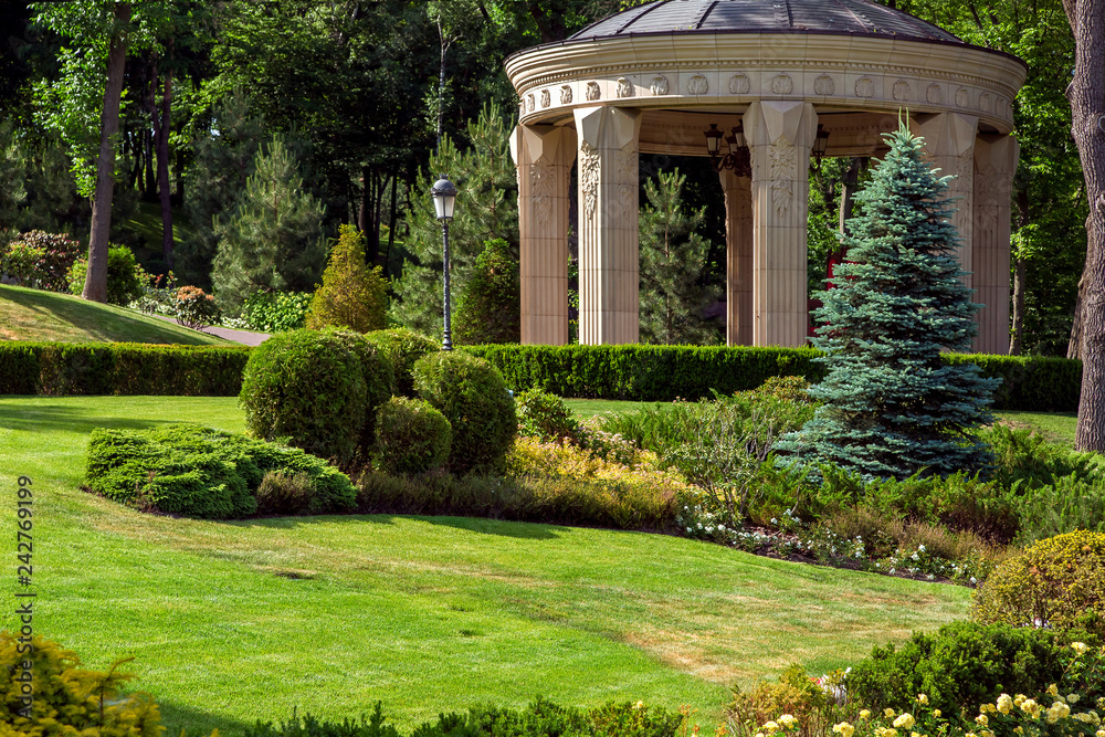 Stone arbor with columns in the park with landscape design and green summer plants in the foreground meadow with lawn and bushes, in the background deciduous trees.