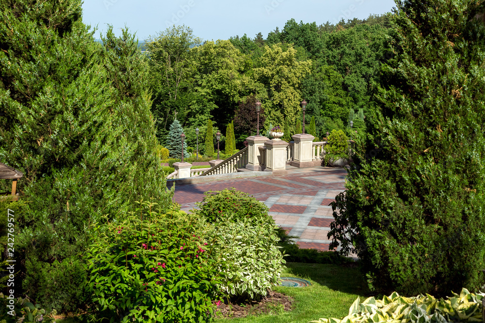 landscaping flower beds with bushes and evergreen thuja in the background a sidewalk with a walkway and a staircase with railings down to the forest.