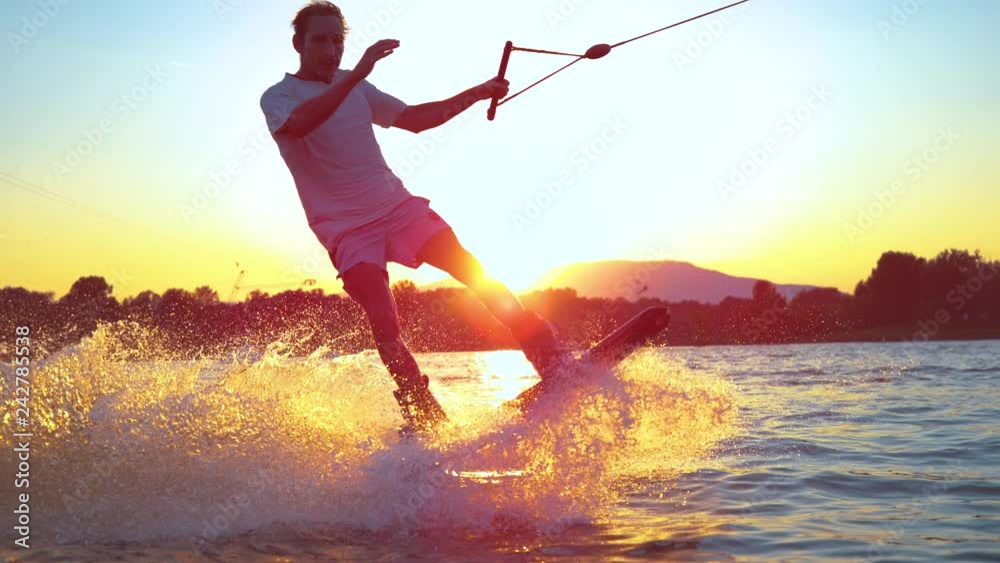 SLOW MOTION CLOSE UP: Surfer wakeboard jumping 180 ollie at golden sunset. Skilled wakeboarder doing water ollie jump, splashing water drops into camera. Young man riding cable wake park on vacation