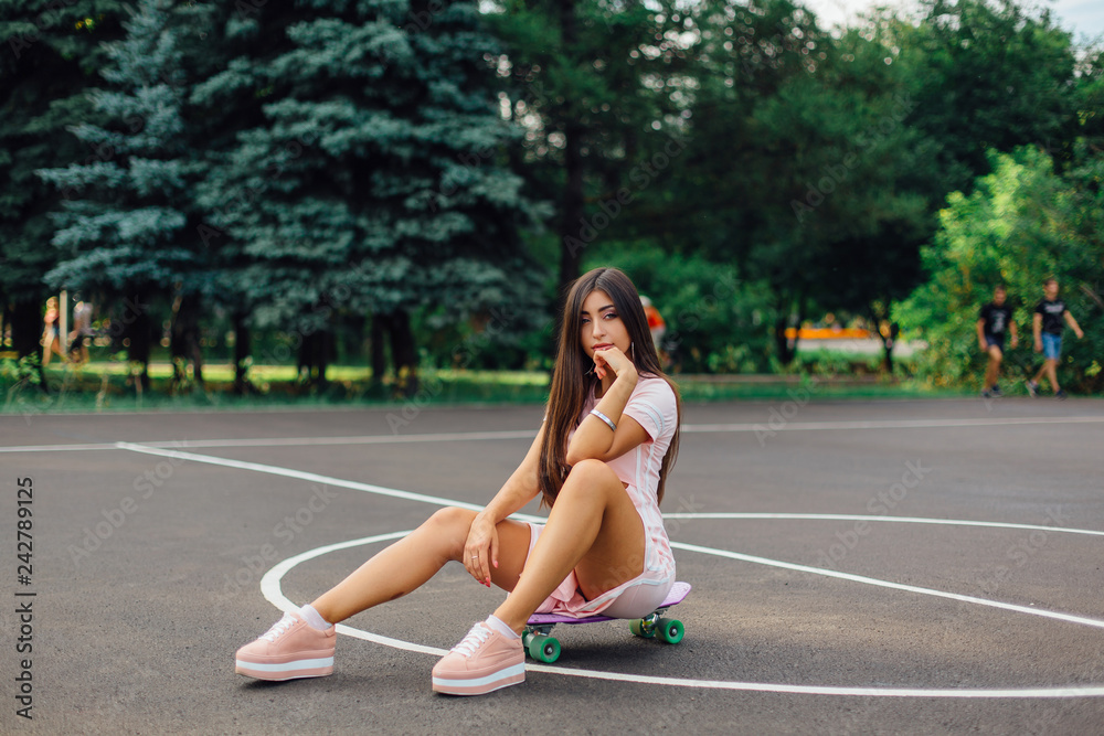 Portrait of a smiling charming brunette female sitting on her skateboard on a basketball court. Portrait of a smiling charming brunette female sitting on her skateboard on a basketball court.