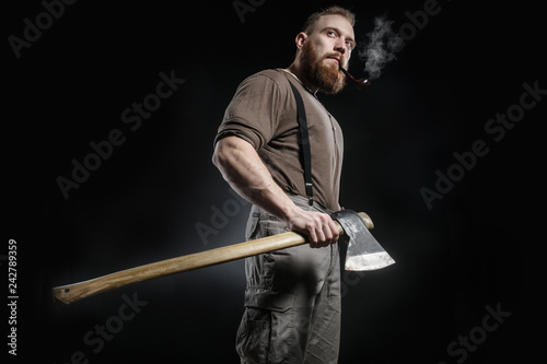 Lumberjack brutal bearded muscled man in brown shirt with smoking tube and suspenders on grey pants and axe on dark background