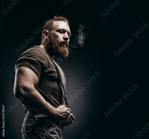 Lumberjack brutal red beard muscled man in brown shirt with smoking tube standing on dark background. Handsome man with red beard and moustache smoking pipe photo