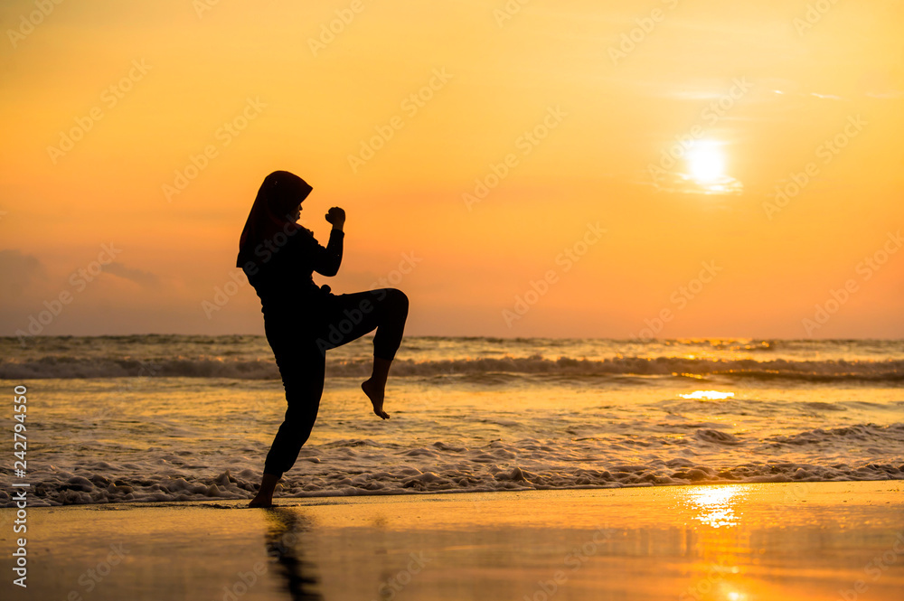 silhouette of young fit Muslim woman covered in Islam hijab head scarf training martial arts karate kick attack and fitness workout at beautiful beach sunset