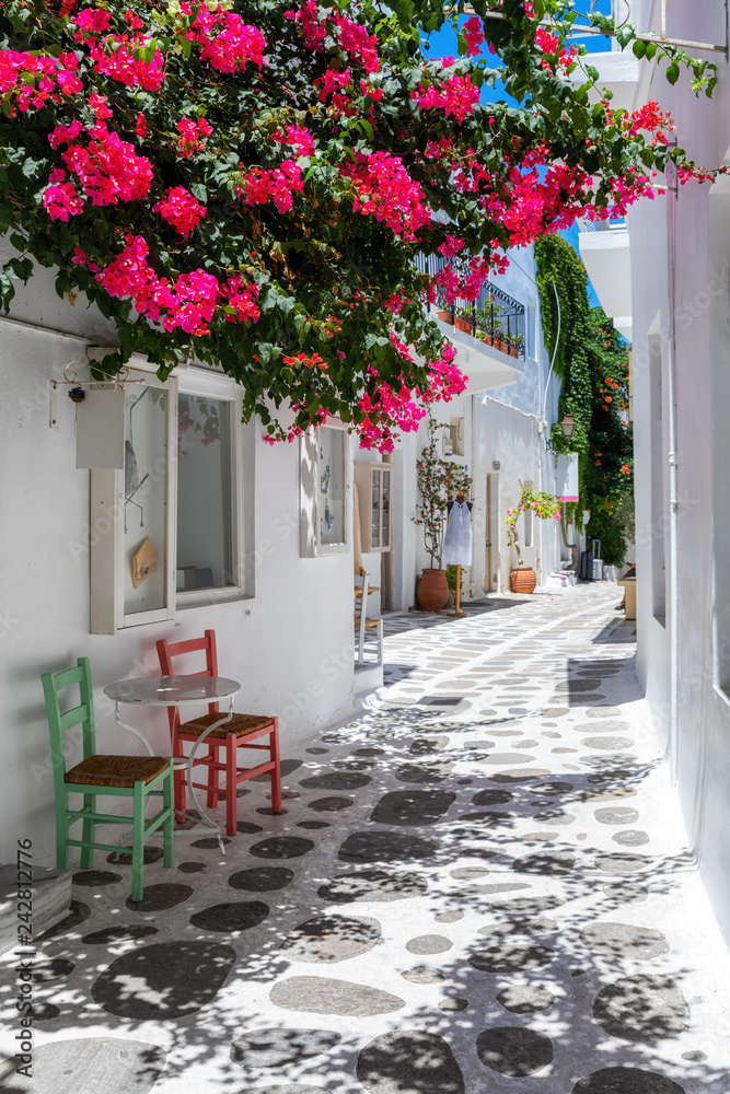 Weiße Gasse mit bunten Bougainvillea Blumen, weißen Häusern und farbigen Stühlen auf den  Kykladen in Griechenland  - obrazy, fototapety, plakaty 