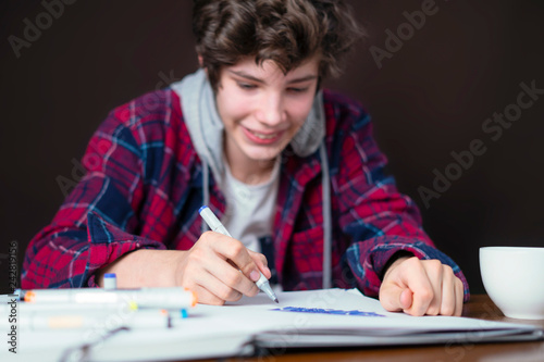 young male sitting at the table and drawing pictures in his notebook with marker b