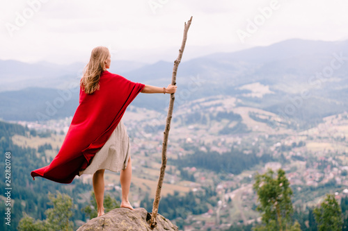 Unknown female in dress and red cape standing on stone at top of mountaing. Barefoot blonde woman with  big wooden stick enjoying landscape from point of view. Girl in red cloak outdoor portrait. photo