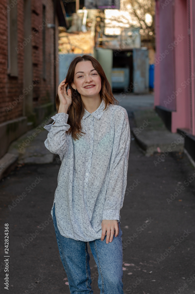 Fototapeta premium young beautiful girl dressed in a lightweight shirt and light blue jeans poses for a photo in the middle of the street, smiling