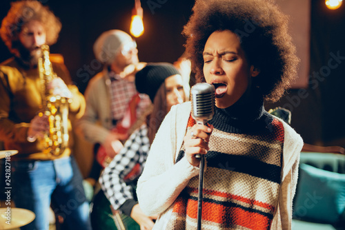 Close up of mixed race woman singing. In background band playing instruments. Home studio interior.