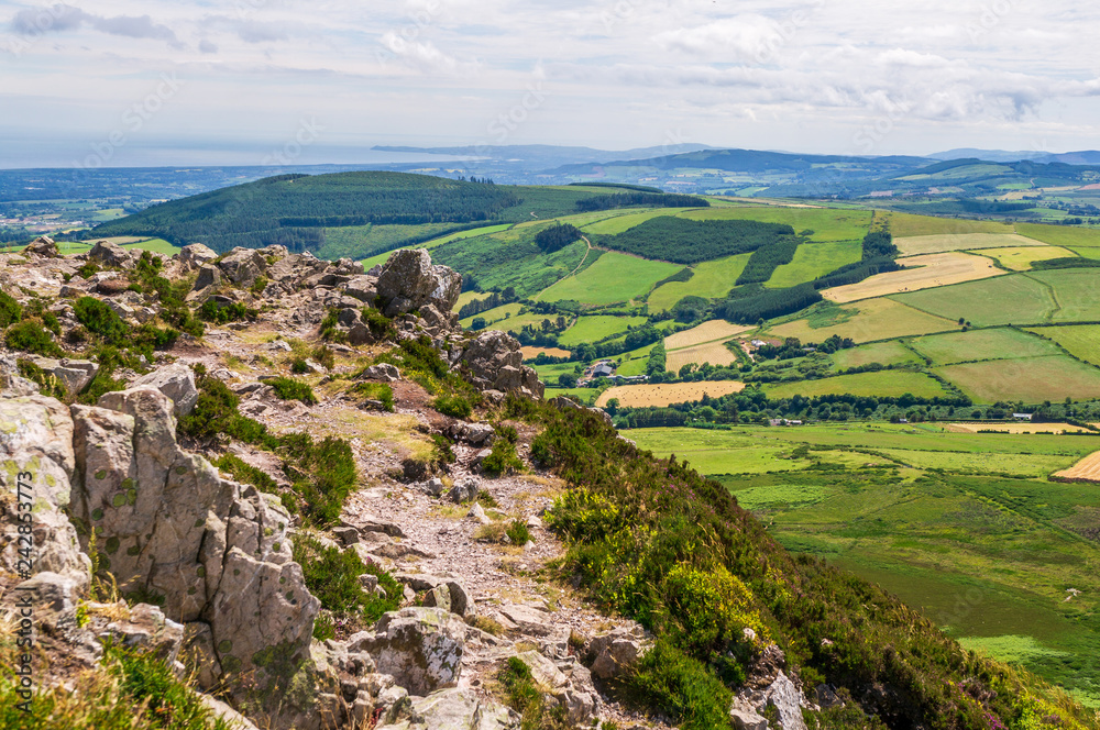 Mountain peak landscape with rugged rocks and distant hills covered in different shades of green. View from the top of The Great Sugar Loaf Mountain in Wicklow, Ireland on a sunny Summer day.