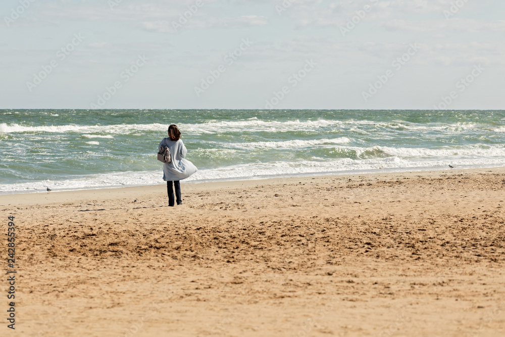 Young woman (brunette) in a light blue cardigan and navy jeans, with a striped backpack, walks along the beach. Young woman (brunette) in a light blue cardigan and navy jeans, with a striped backpack, walks along the beach.