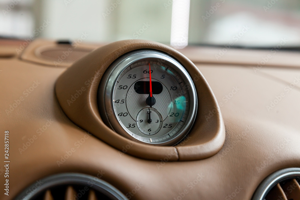 The watch on the dashboard of an expensive car is close-up, covered with natural beige leather, with a white dial, a red arrow and a metal chrome rim.