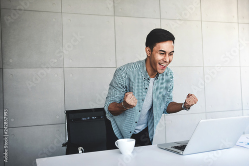 Small Business and Successful Concept. Young Asian Businessman Glad to recieve a Good News or High Profits from Computer Laptop, Own Business Achieves Goals. Raising Arms to Celebrating Success