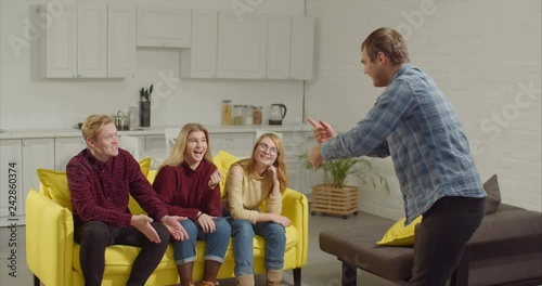 Cheerful man acting out a word in the game of charades, gesturing and showing pantomime to engrossed diverse teenage friends at home. Smart friends guessing word in charades while relaxing indoors.