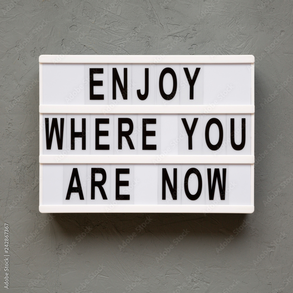 'Enjoy where you are now' words on lightbox over concrete background, top view. Flat lay, from above, overhead.