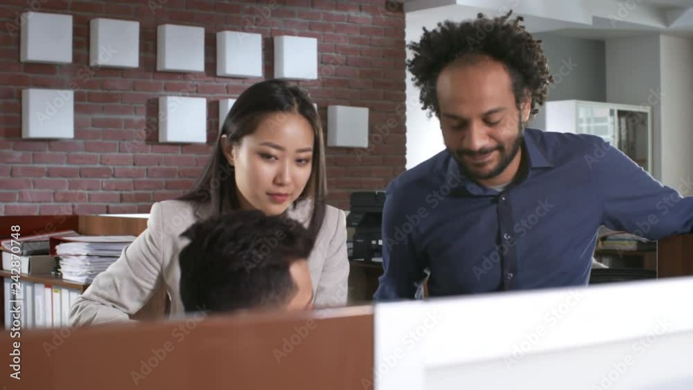 Group of three multi ethnic colleagues discussing work in office, one of them sitting at desk and the other two standing near him Group of three multi ethnic colleagues discussing work in office, one of them sitting at desk and the other two standing near him