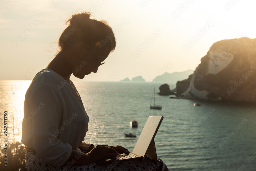 Young woman using laptop computer at sunset in front of the sea on Ponza island coast, sitting on a wall with view of the ocean.