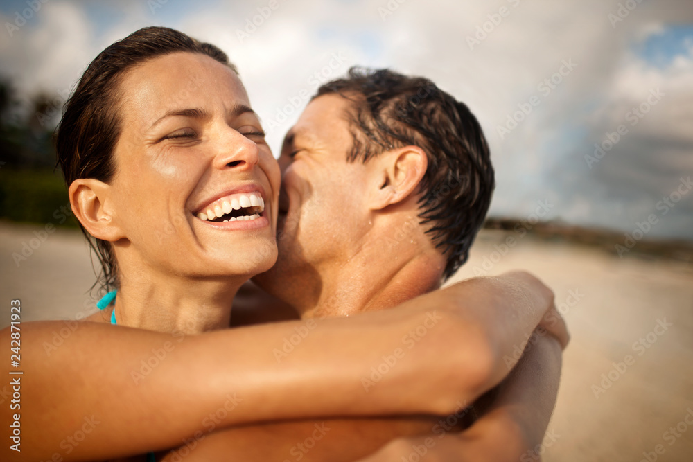 Smiling mid-adult woman being kissed on the cheek by her husband on a beach. Smiling mid-adult woman being kissed on the cheek by her husband on a beach.