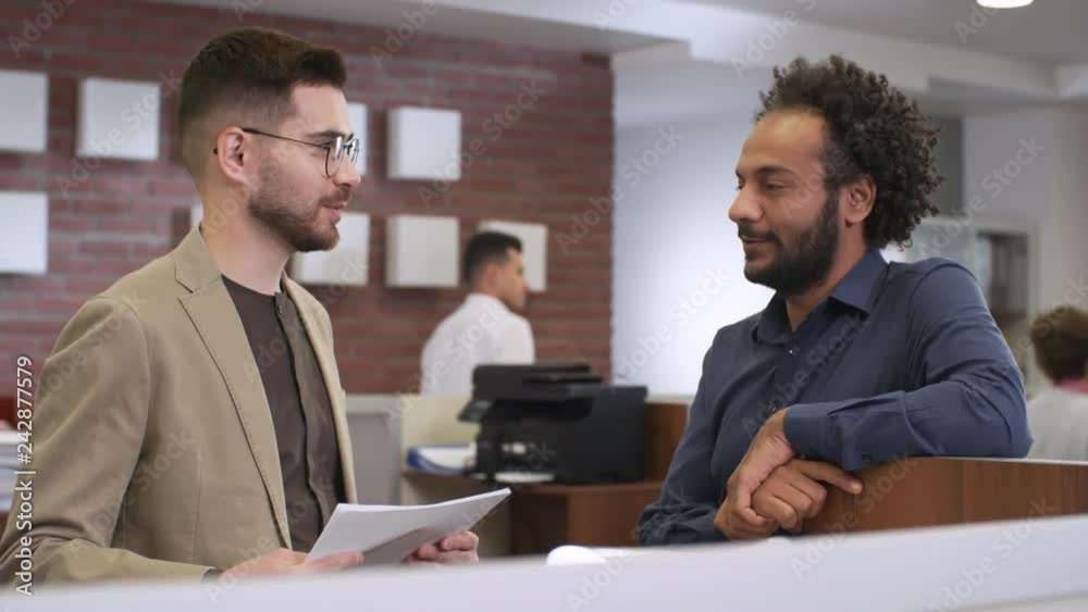 Medium shot of two young men of different ethnicities talking to each other while taking break from work in office