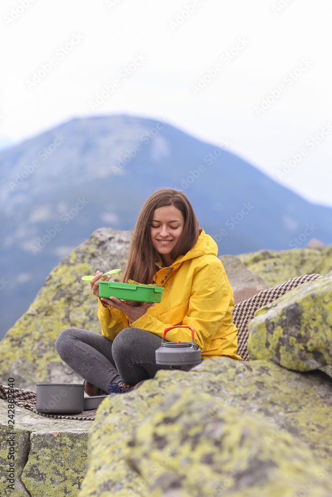 Young woman in yellow jacket have a picnic on top of the mountain. 