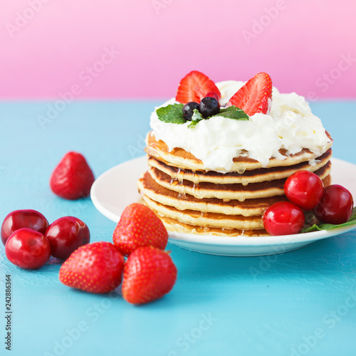 pancakes in a plate with whipped cream, honey, strawberries, mint and sweet cherries on a bright blue and pink background.