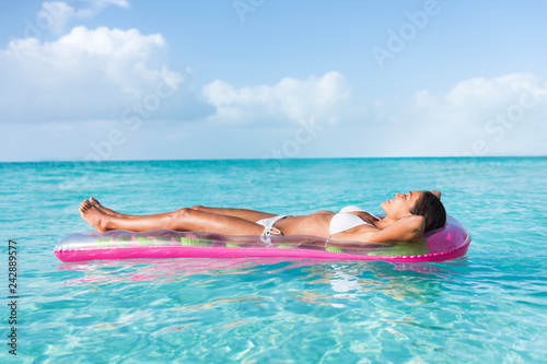 Luxury summer vacation beach woman relaxing lying down on inflatable pool float floating on turquoise ocean sun tanning. Bikini model sleeping on holiday.