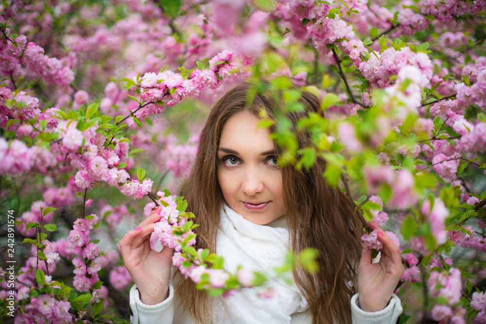 .A beautiful fair-haired girl with green eyes in a white coat and a white scarf stands in the forest in a pink cherry blossom and holds branches with her hands. Horizontal photography. .A beautiful fair-haired girl with green eyes in a white coat and a white scarf stands in the forest in a pink cherry blossom and holds branches with her hands. Horizontal photography.