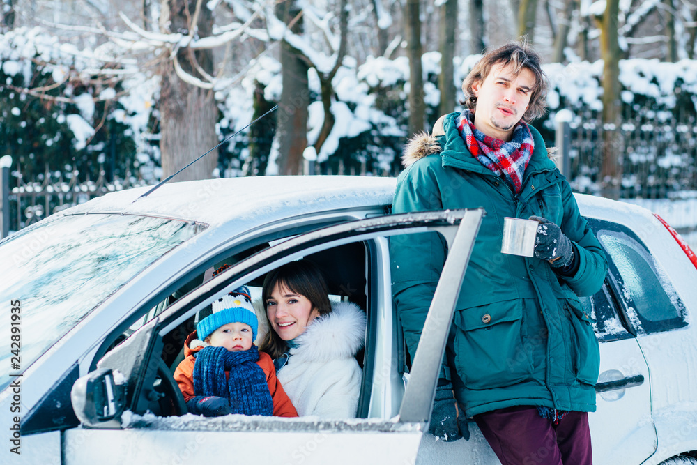 Family of three, dad, mom and toddler son travel by car in cold winter season. Father standing behind car outdoor and opening thermos for warm drink.