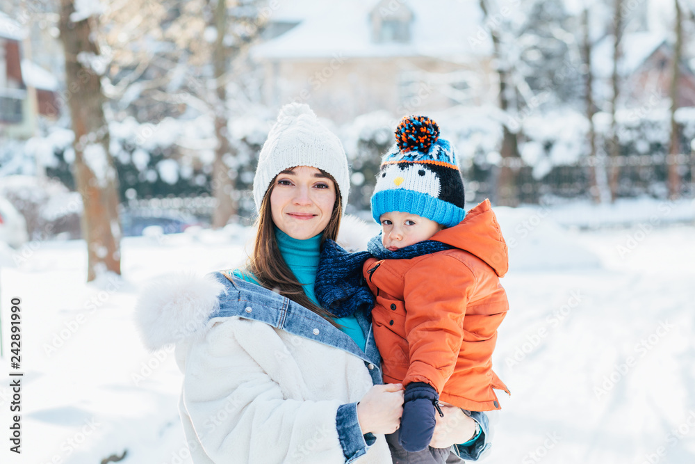 Cheerful woman in white jacket holding her sad little toddler son on her arms outdoor in winter day. Holidays, people, family, fun and vacation concept.