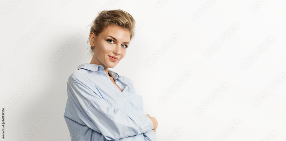 Portrait of young female wearing light blue shirt and standing with arms crossed. Elegant model with kind smile and beautiful nude makeup. Isolated on white background