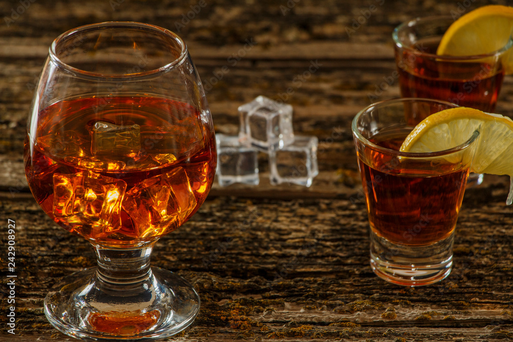 Whiskey with ice or brandy in a glass and a square carafe on an old wooden background. Whiskey with ice in a glass. Whiskey or cognac. Selective focus. Whiskey with ice or brandy in a glass and a square carafe on an old wooden background. Whiskey with ice in a glass. Whiskey or cognac. Selective focus.
