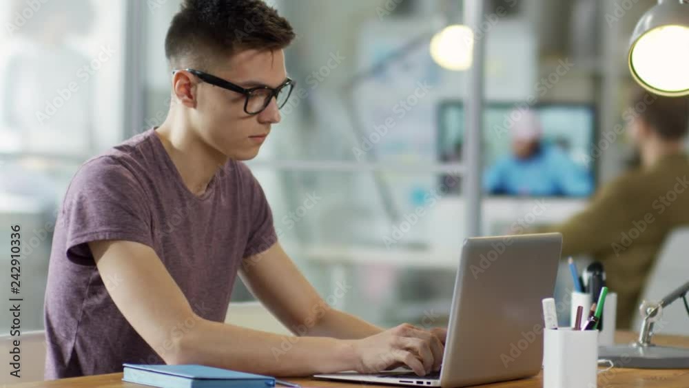 Tracking shot medium shot of young male office worker in tshirt and glasses sitting at office desk and typing on laptop computer